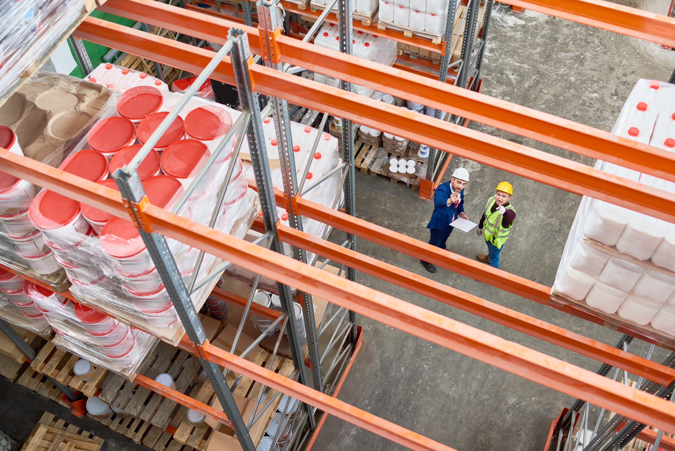 Top view background image of tall shelves in modern warehouse with two workers wearing hardhats standing in aisle and pointing up